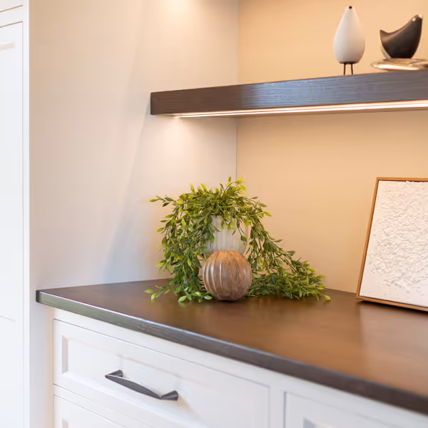 Modern kitchen shelving with wooden floating shelves displaying glassware, a small framed photo, bowls, a decanter, and flowers, above a countertop and wooden cabinets, with a dining table and chairs in the foreground.