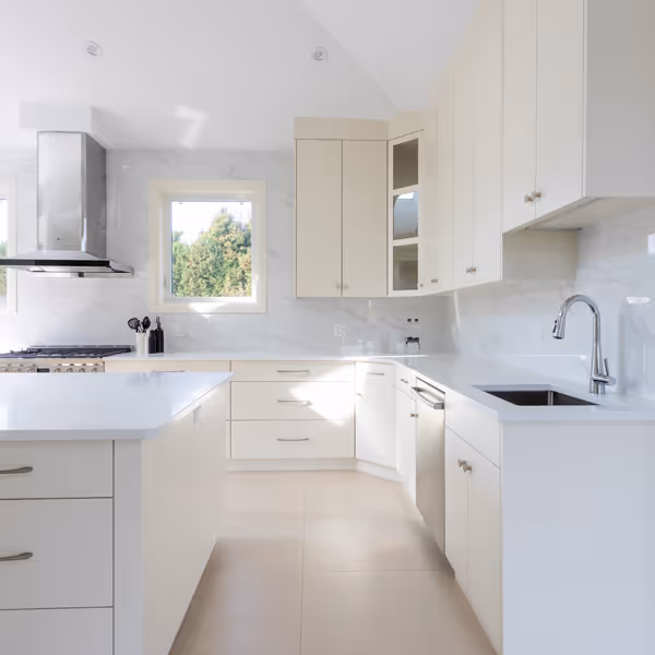 Modern white kitchen with stainless steel refrigerator, built-in oven and microwave, hardwood floor, and a woven chair at a white countertop.