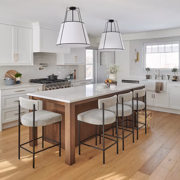 Modern white kitchen with stainless steel refrigerator, built-in oven and microwave, hardwood floor, and a woven chair at a white countertop.
