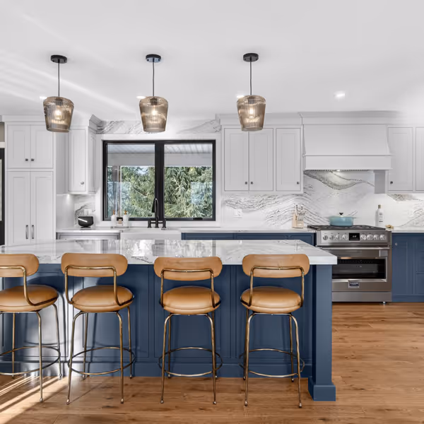 Modern white kitchen with stainless steel refrigerator, built-in oven and microwave, hardwood floor, and a woven chair at a white countertop.