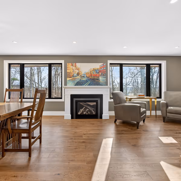Cozy living room with a white fireplace decorated with greenery and red roses, a mounted flat-screen TV above it, a leather sofa, framed artwork on the walls, glass coffee table, and soft lighting.