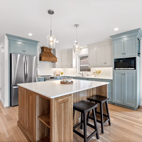 Modern white kitchen with stainless steel refrigerator, built-in oven and microwave, hardwood floor, and a woven chair at a white countertop.
