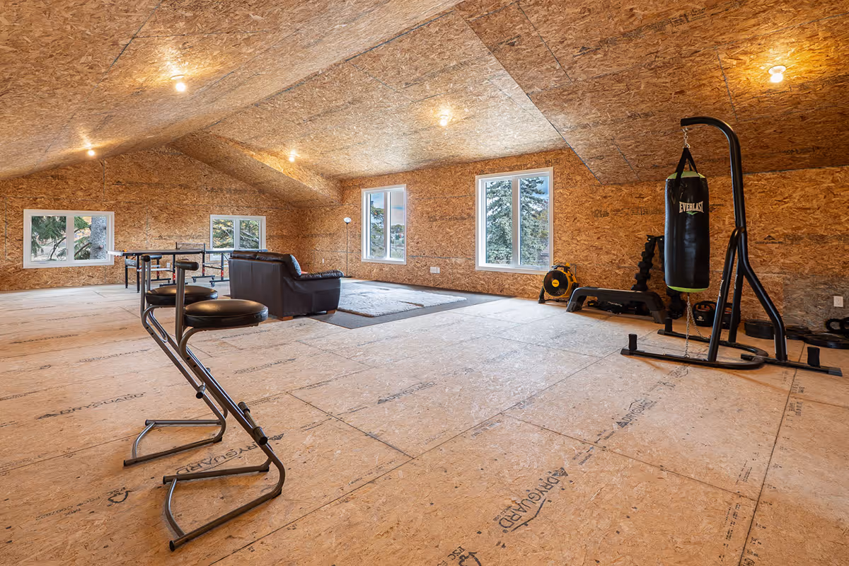 Modern basement living area with beige sofas, black round coffee table, wall-mounted TV, and exercise equipment in the background.