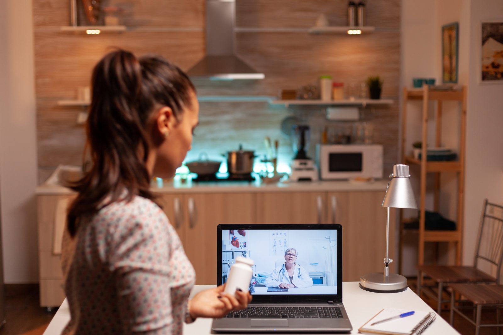 Woman at her kitchen table holding up a medication bottle during a video consultation with a doctor on her laptop