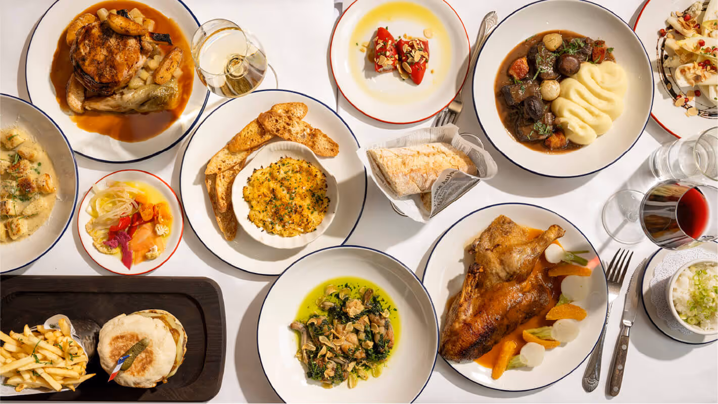 Overhead view of a table spread with various plated gourmet dishes including roasted chicken, mashed potatoes, grilled sandwich with fries, pickled vegetables, and glasses of white and red wine.