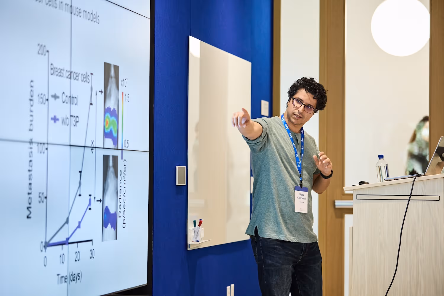 Man in glasses and lanyard giving a presentation pointing at a screen showing a scientific graph about tumor burden over time.