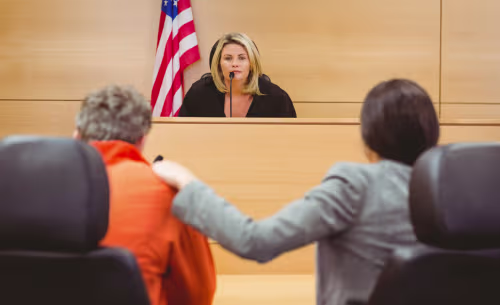 A judge presides over a courtroom hearing while a defendant and defense attorney appear before the bench during a criminal court proceeding.