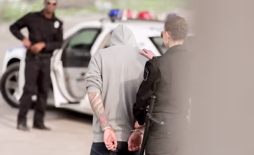 A person in handcuffs is escorted toward a police car by a law enforcement officer, illustrating the initial step of a DUI arrest before booking records can be used to locate them.
