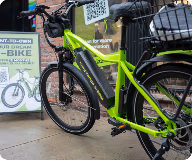 Green electric bicycle with a basket parked on pavement next to a promotional sign about rent-to-own e-bikes.