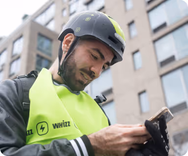 A man wearing a black helmet and bright green Whizz safety vest looking at his phone outdoors in an urban setting.