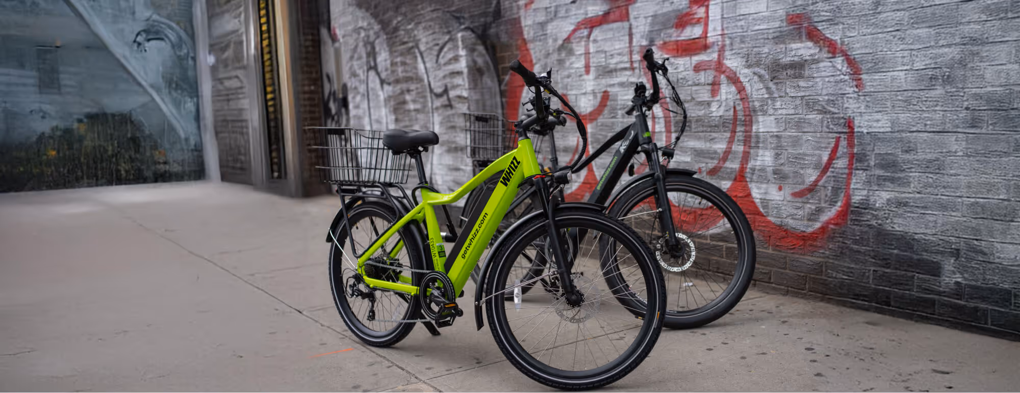 Two electric bicycles, one bright green and one black, parked on a sidewalk in front of a graffiti-covered brick wall.