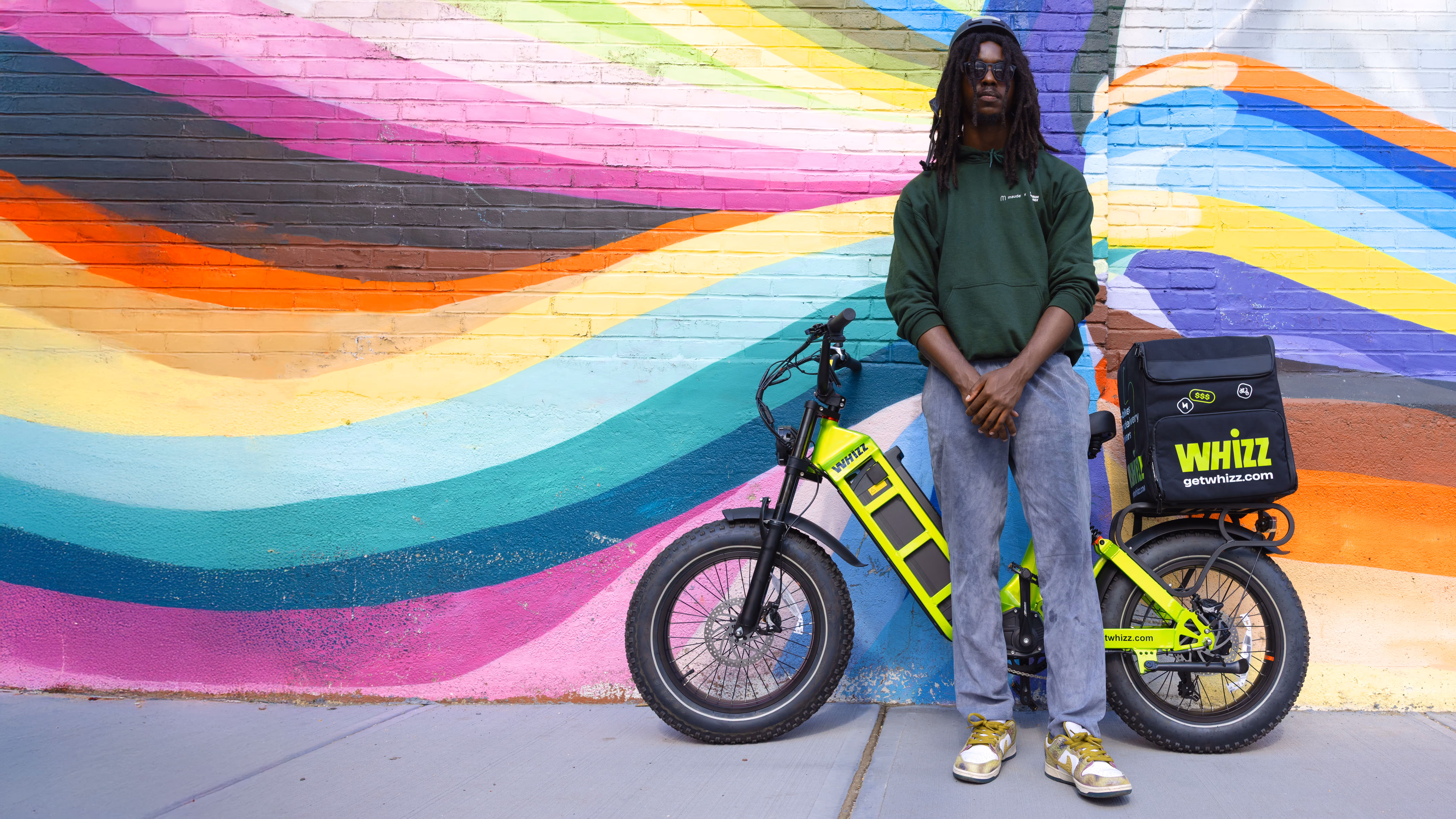 Person in green hoodie and jeans standing next to a bright yellow Whizz delivery electric bike in front of a colorful mural wall.