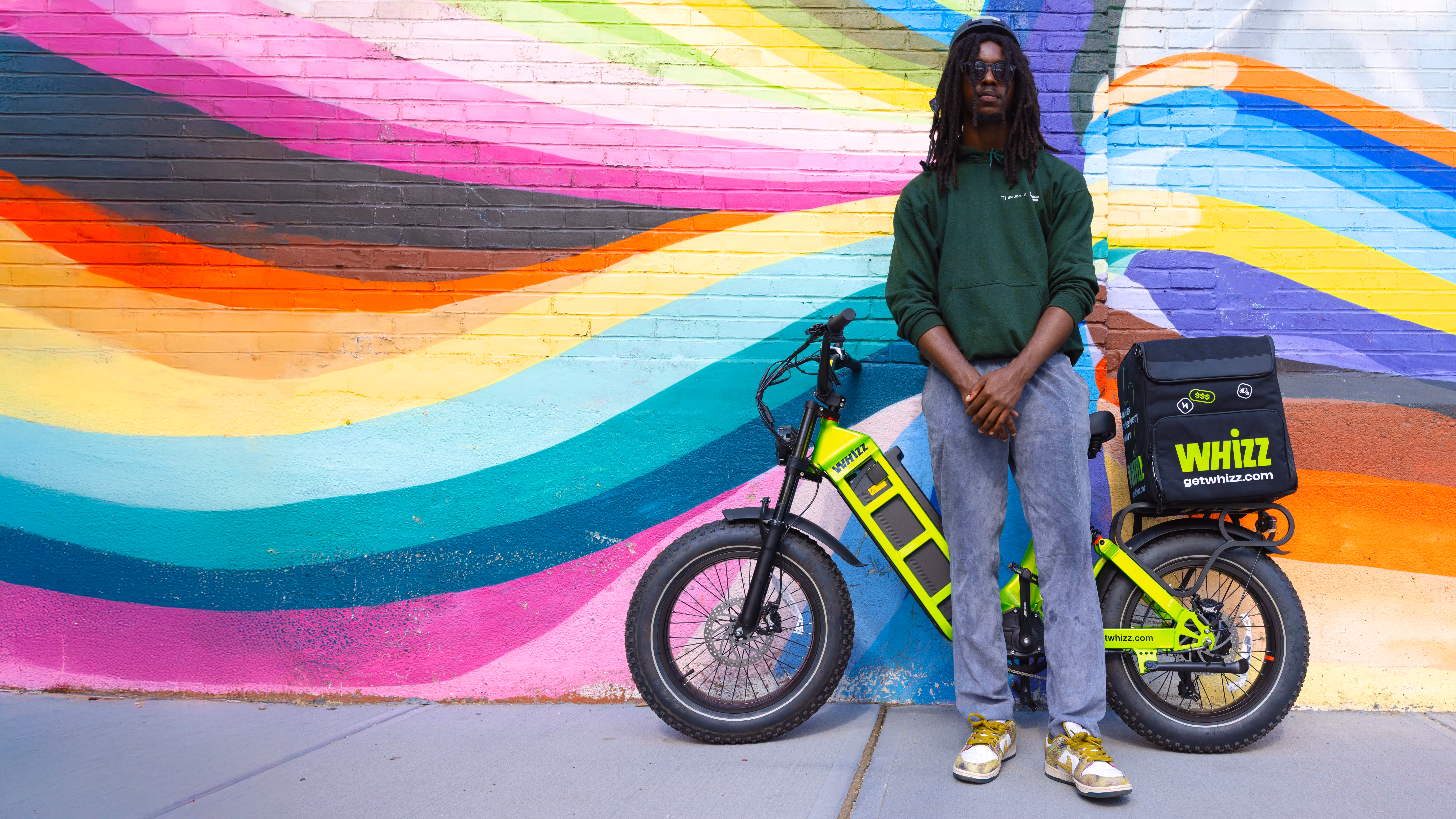 Man wearing a green hoodie and sunglasses standing with a bright yellow Whizz delivery electric bike in front of a colorful mural wall.