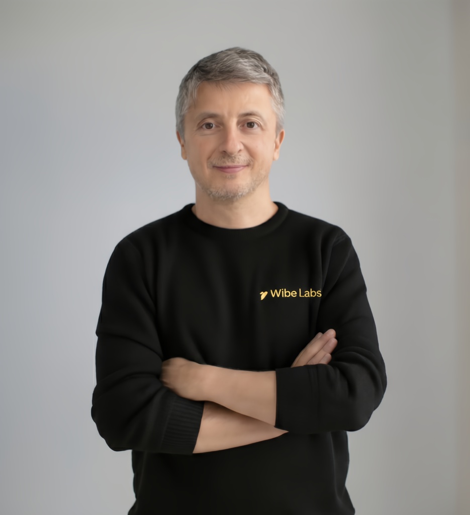 Middle-aged man with short gray hair wearing a black Wibe Labs sweatshirt, standing with arms crossed and smiling slightly against a plain gray background.
