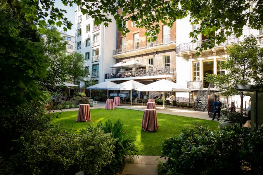A manicured garden with several high-top tables draped in pink cloths, set up on a green lawn in front of a building with outdoor seating and white canopies.