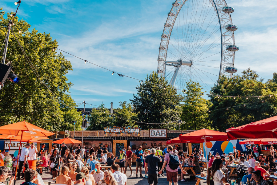 A lively outdoor market with many people dining at picnic tables under red and orange umbrellas, with the London Eye visible in the background on a sunny day.