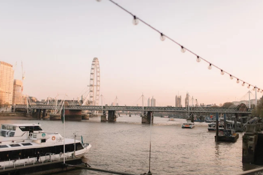 A wide view of the River Thames in London with boats on the water and the London Eye and bridges visible in the background under a pale pink sky.