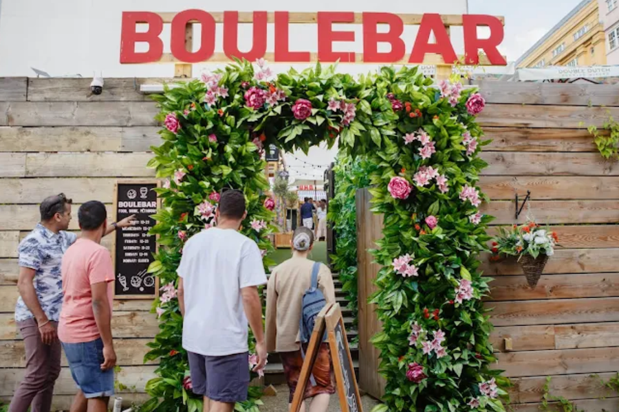 A floral archway leads to an entrance with the word BOULEBAR in large red letters above. People are entering the establishment.