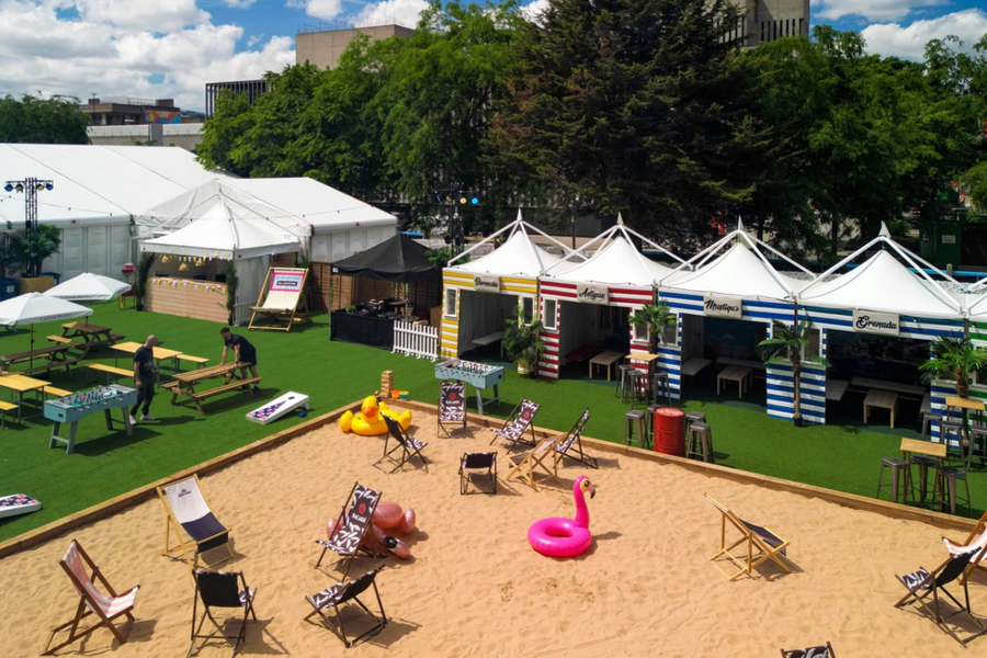 An outdoor event space with a sandy area featuring beach chairs and inflatable toys, adjacent to a green area with picnic tables, foosball, and food stalls under tents.