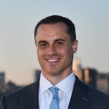 Smiling man in a dark suit and light blue tie standing outdoors with a city skyline in the background.