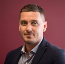 Portrait of a man with short brown hair wearing a dark blazer and light blue shirt against a maroon background.