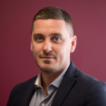 Portrait of a man with short brown hair wearing a dark blazer and light blue shirt against a maroon background.