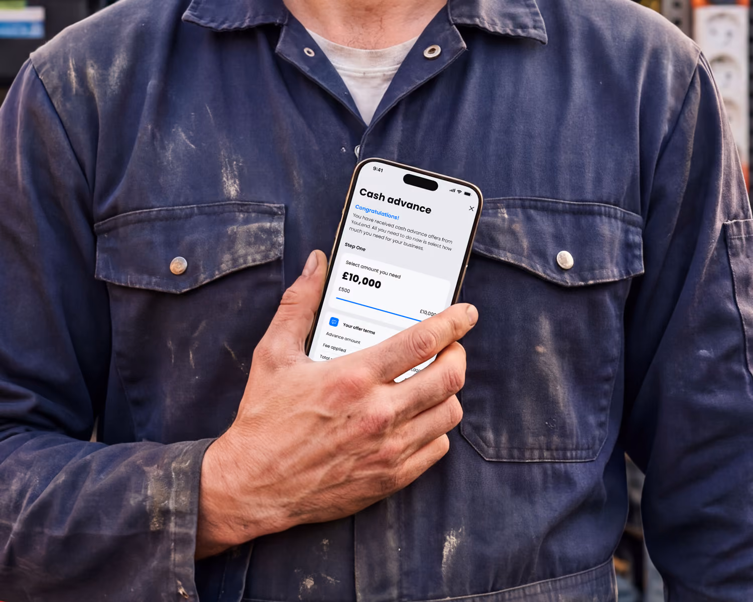 An electrician holding phone displaying a cash advance screen on his phone with £10,000 selected.