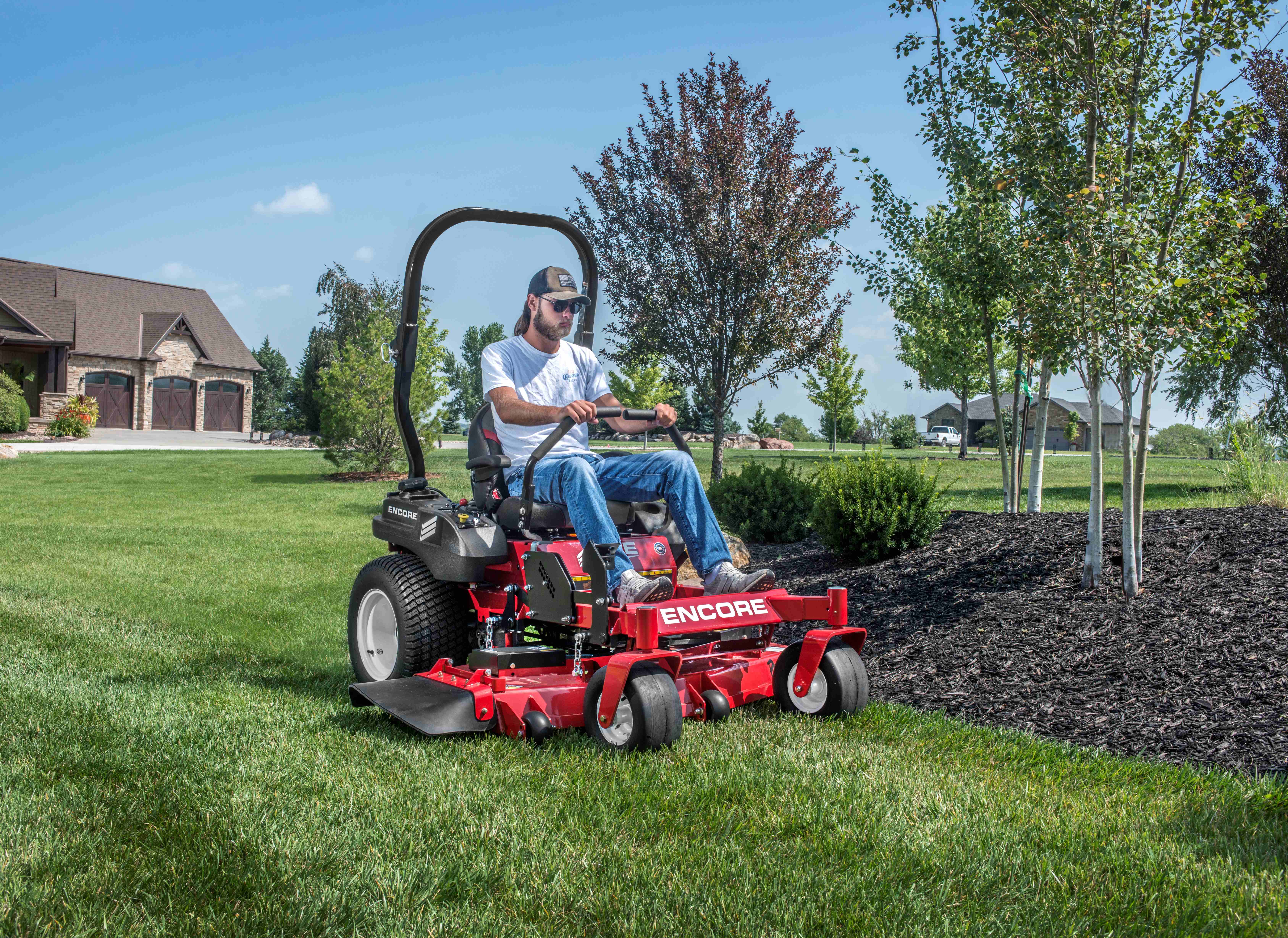 Man wearing sunglasses and cap riding a red Encore zero-turn lawn mower on a green lawn near trees and mulch beds.