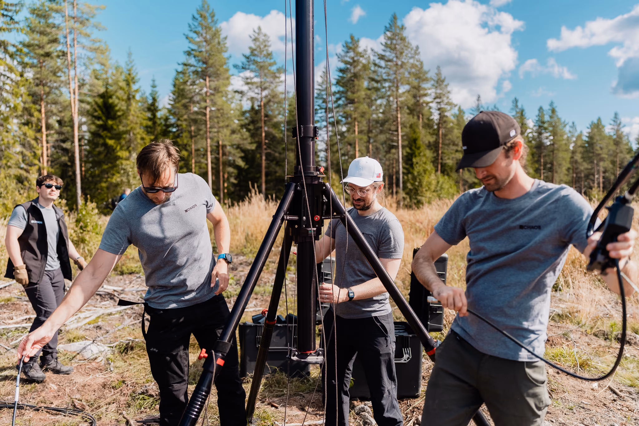 Four crew members in grey CHAOS shirts raising a tripod antenna mast in a pine forest clearing