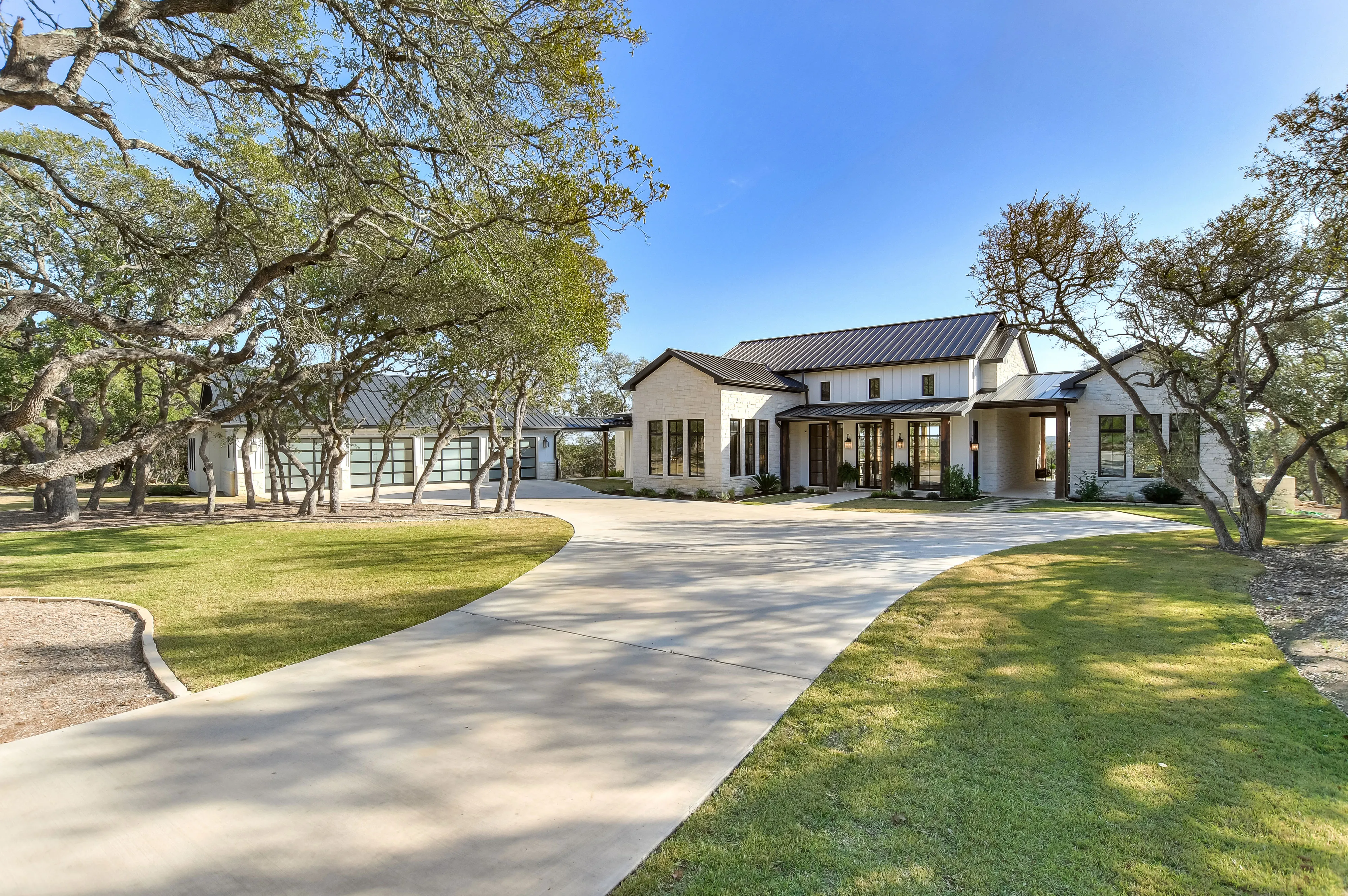 White home with a dark metal roof and a curved concrete drive under live oaks