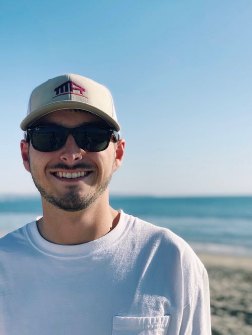 Smiling man wearing sunglasses and a beige cap standing on a beach with ocean in the background.