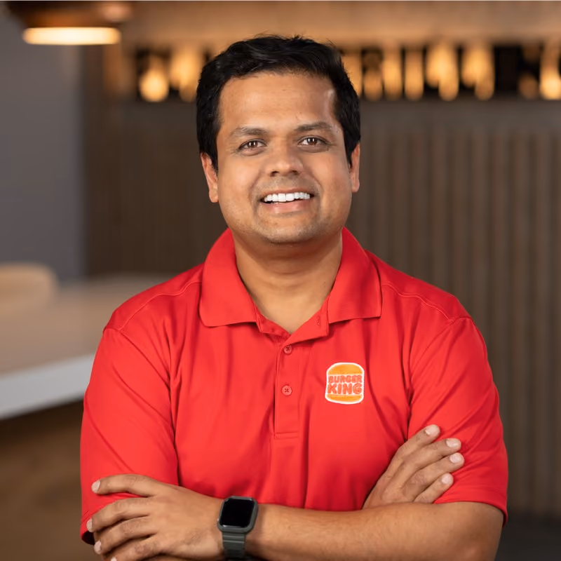 Smiling man wearing a red Burger King polo shirt with arms crossed and a black smartwatch on his left wrist, standing indoors with blurred background.