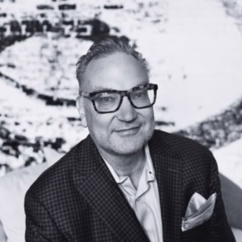 Middle-aged man with slicked-back gray hair and glasses wearing a checkered blazer and light-colored shirt with a pocket square, smiling slightly against a blurred abstract background.