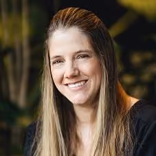 Smiling woman with long straight light brown hair wearing a black top, sitting indoors with blurred plants in the background.