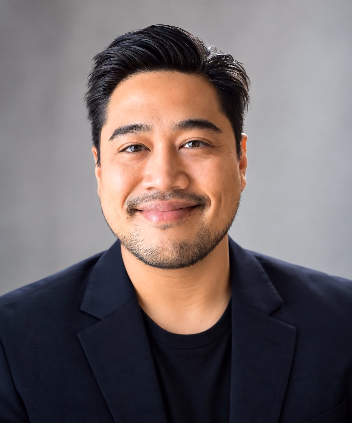 Portrait of a smiling man with neatly styled black hair and light facial hair, wearing a black blazer over a black shirt, against a neutral gray background.