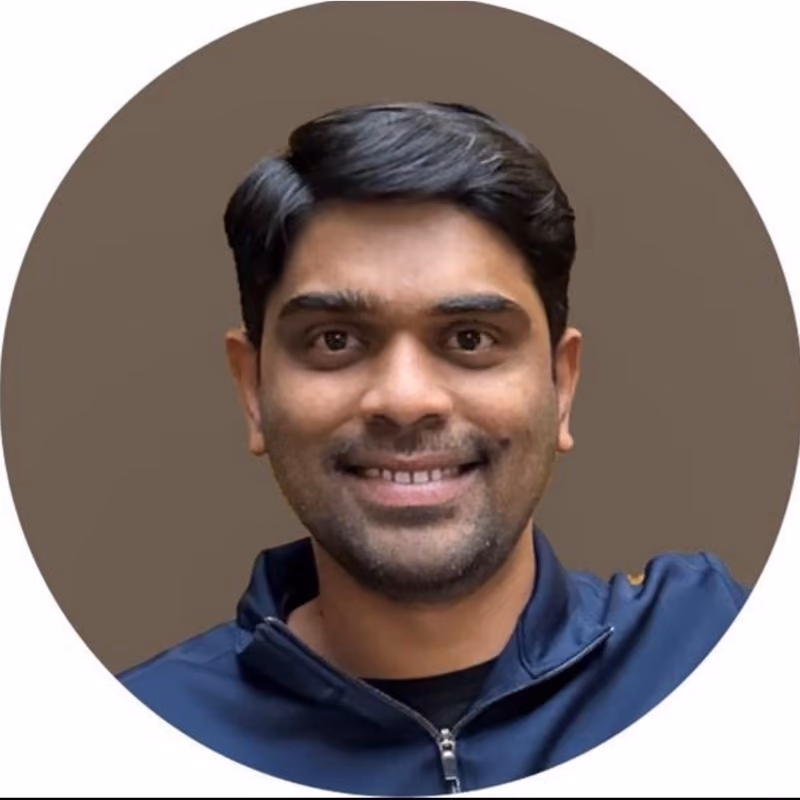 Portrait of a smiling man with short black hair, wearing a dark blue zip-up jacket, on a plain brown circular background.