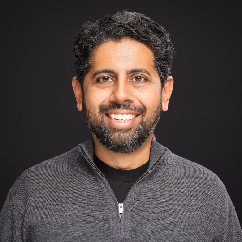 Smiling man with dark curly hair and beard wearing a gray zip-up sweater over a black shirt against a black background.