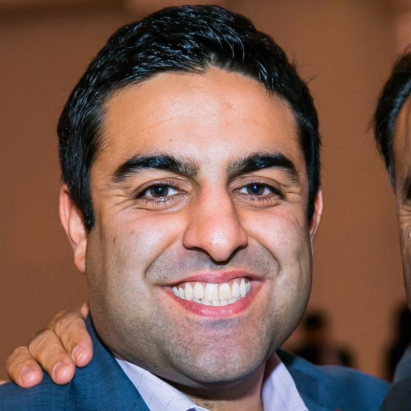 Close-up of a man with dark hair smiling broadly, wearing a blue suit jacket and a light-colored shirt, with a hand resting on his shoulder.