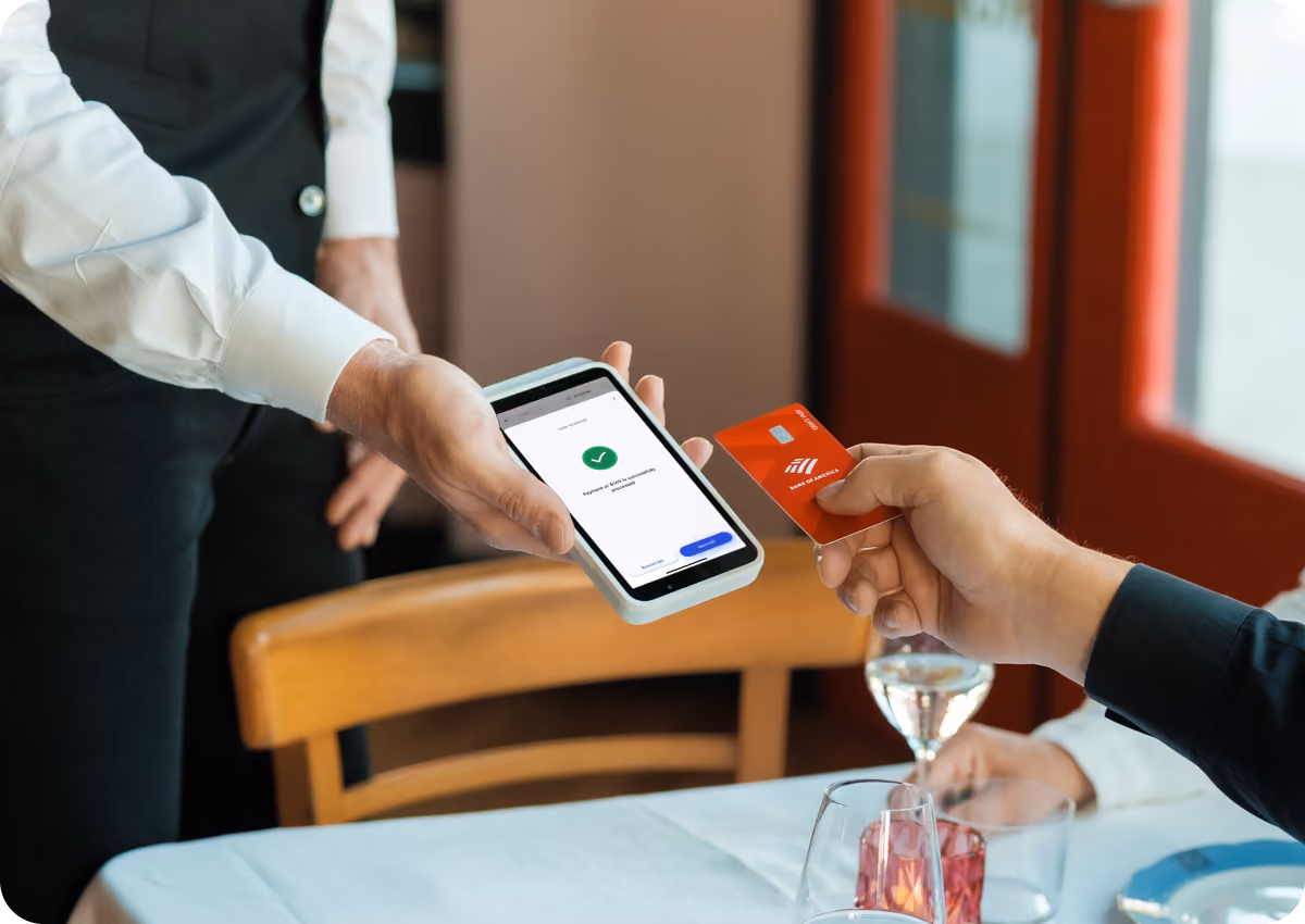 A customer holding a red credit card and making a contactless payment to a waiter holding a point-of-sale device displaying a payment approval screen with a green checkmark, at a restaurant table set with glasses and plates.