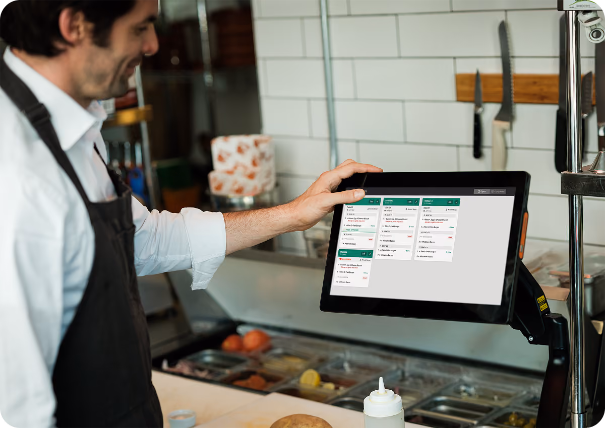 A man wearing a dark apron and white shirt interacts with a kitchen order management screen mounted near a food prep station with ingredients in containers below.