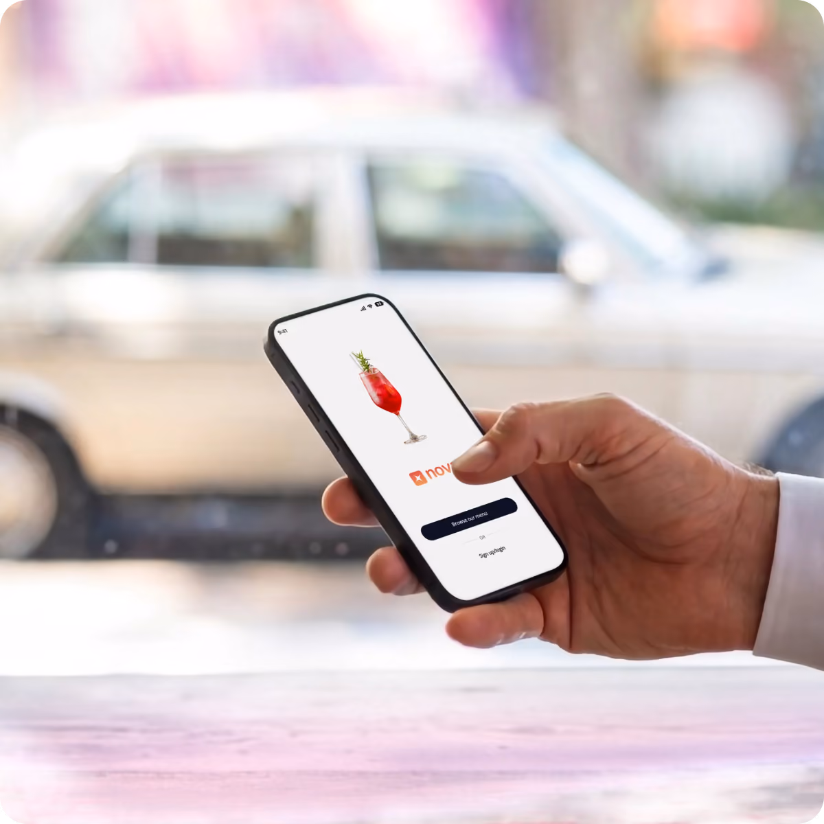A hand holding a smartphone displaying a white screen with a red cocktail glass image and app interface buttons, with a blurred car in the background.