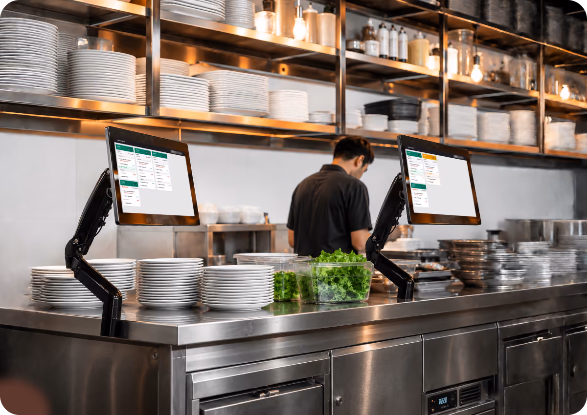Modern commercial kitchen workspace with stainless steel counters holding stacks of white plates, two mounted touchscreen monitors displaying kitchen order systems, and a worker in a black shirt preparing food with containers of fresh green lettuce.