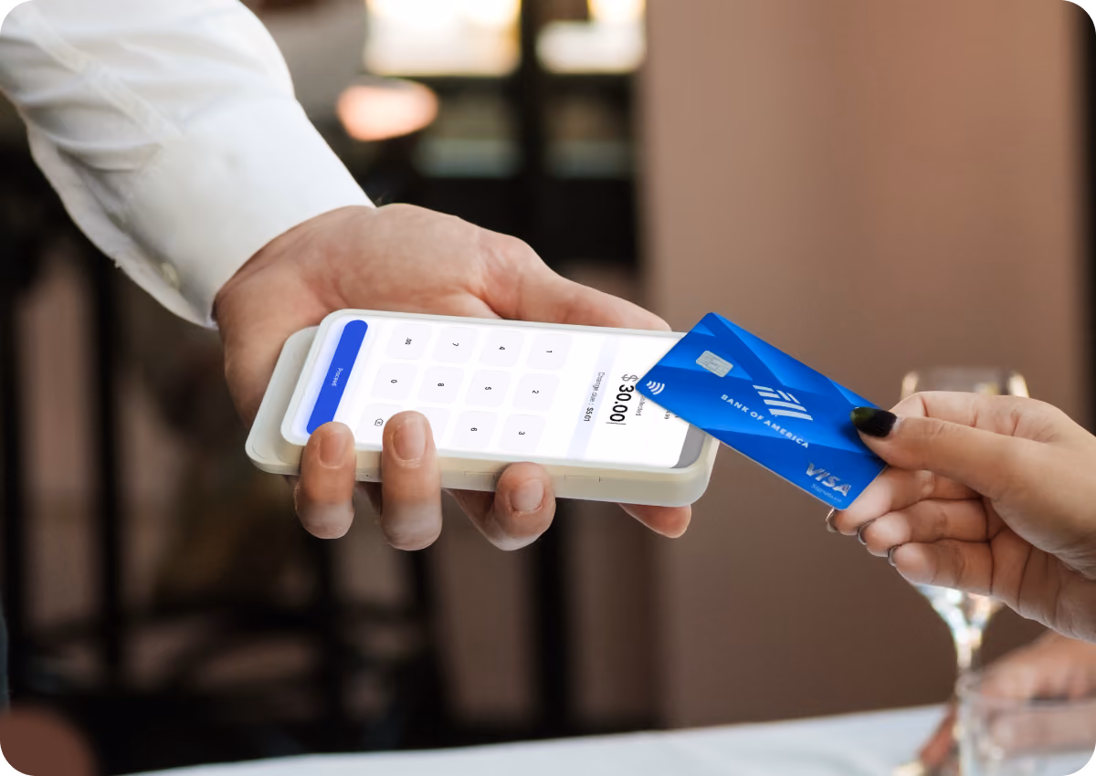 A person holding a white digital card reader showing an input screen with $30.00 displayed, while another hand holds a blue Bank of America Visa card near the reader for contactless payment.