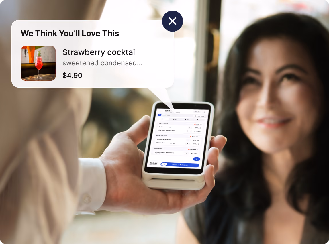 A person holding a smartphone displaying an order screen with a list of food items and prices, while a woman smiles in the background. A popup bubble shows a recommendation for a strawberry cocktail priced at $4.90, with a small image of the drink.