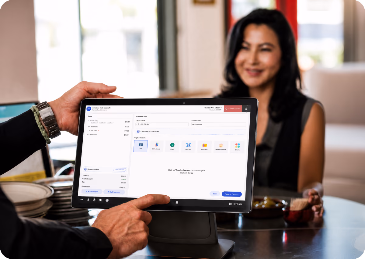 Tablet point-of-sale system screen showing payment mode options including card, cash, and phone account, held by a person with a woman sitting in the background smiling.