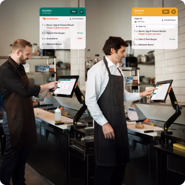 Two men in black aprons using touch screen order systems in a restaurant kitchen with digital order details displayed above each screen.