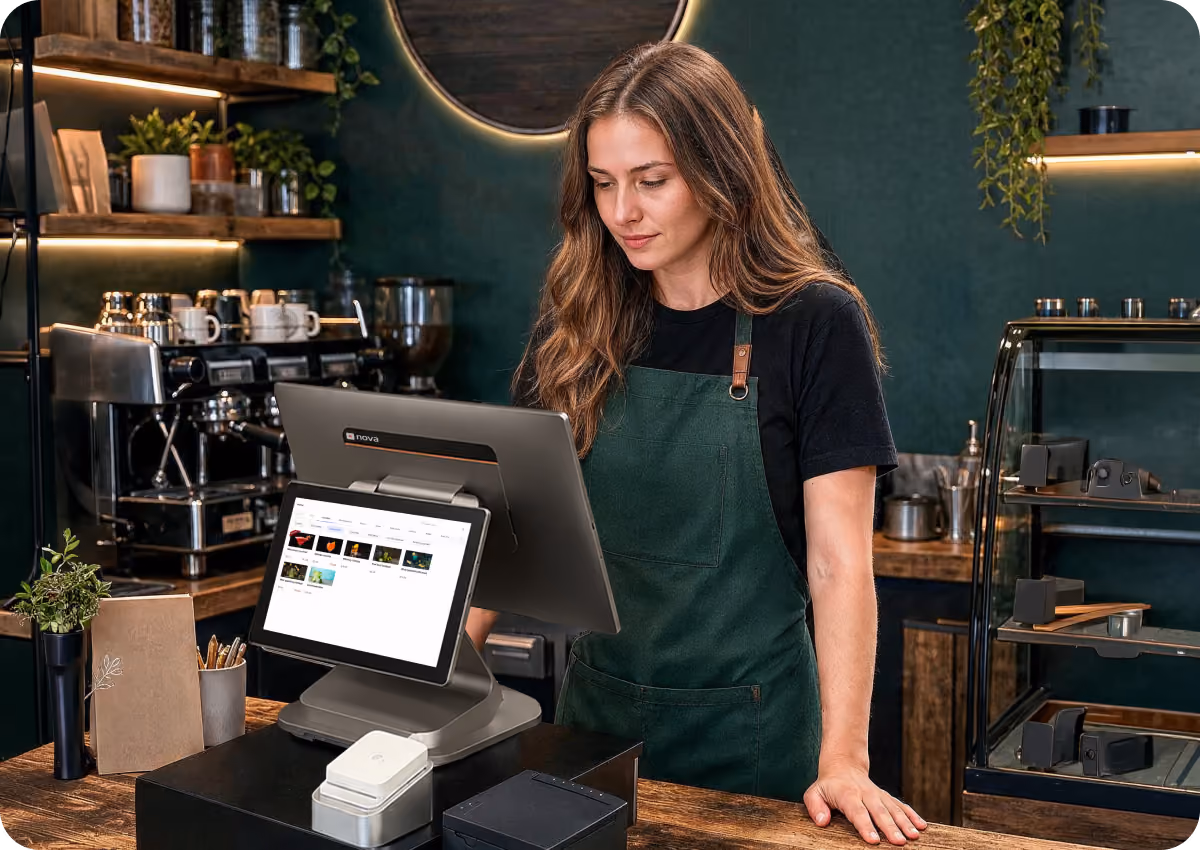 A woman in a green apron and black shirt stands behind a coffee shop counter looking at a point-of-sale touchscreen, with an espresso machine and shelves in the background.