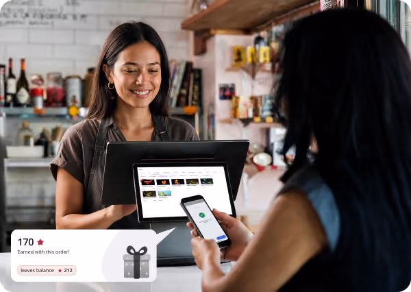 Smiling cashier wearing an apron standing behind a counter with a digital register while a customer uses a smartphone for payment, shown with an overlay indicating 170 points earned from the order and an available balance of 212 points.