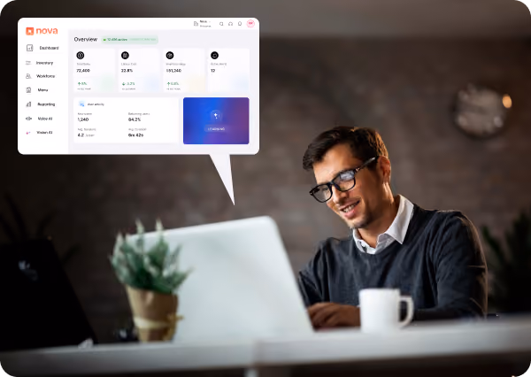 Man with glasses sitting at a desk working on a laptop, smiling, with a coffee cup and a small potted plant on the desk, and a digital interface overlay showing an overview dashboard with metrics and menu options.