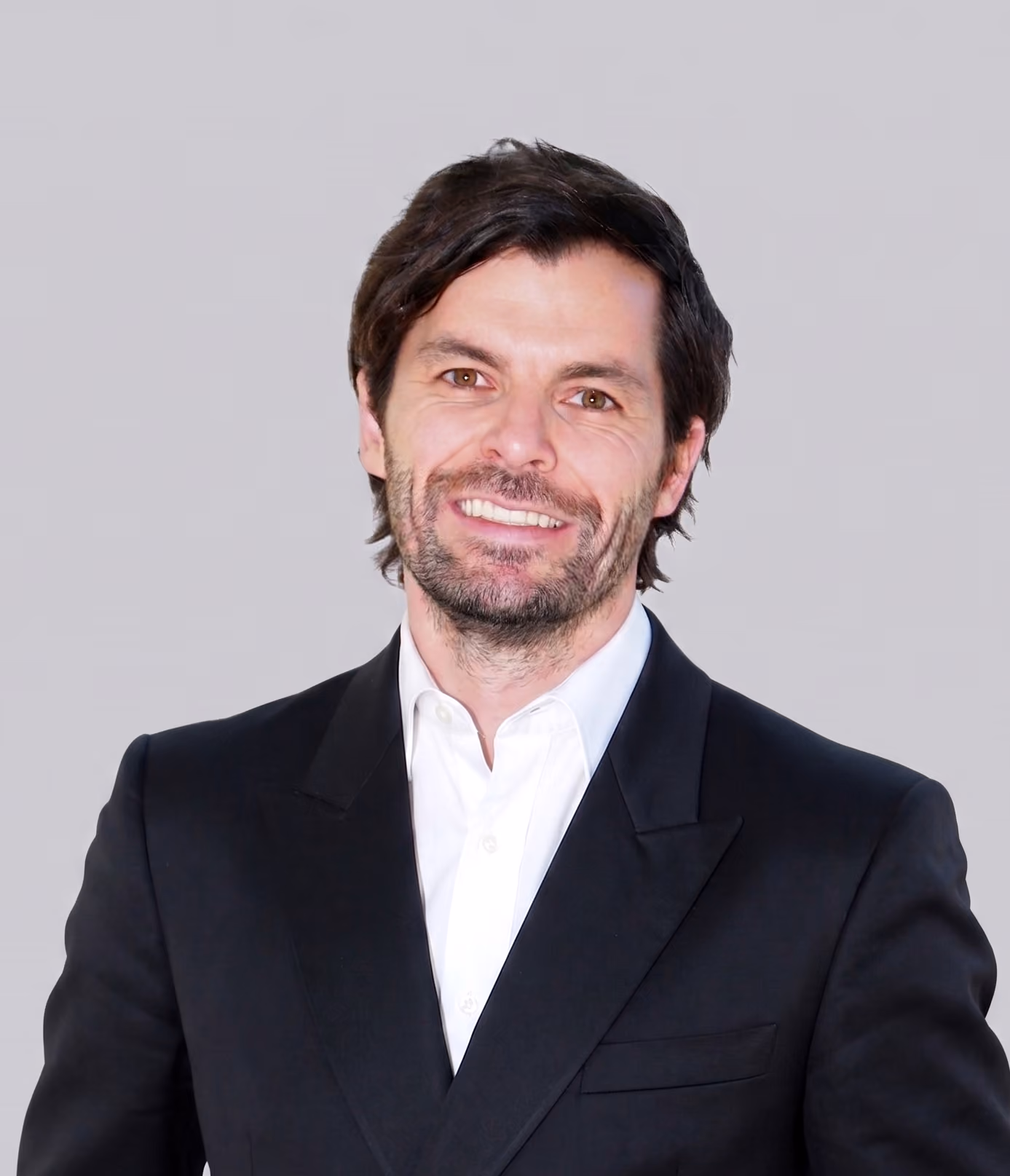 Man with dark hair and beard wearing a black suit jacket and white shirt, smiling against a plain gray background.